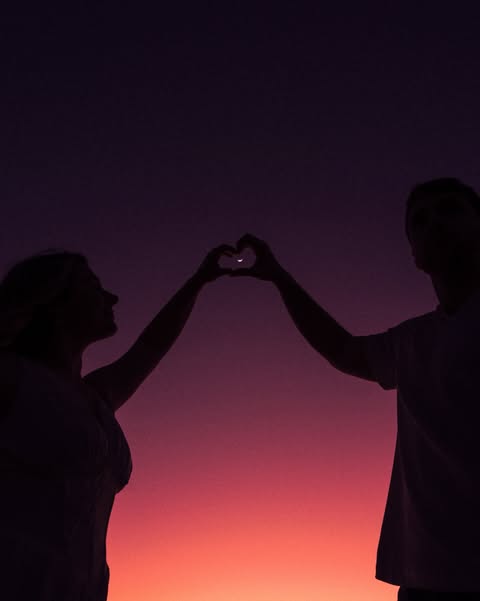 A couple joining their hands into a heart against a violet and orange sky