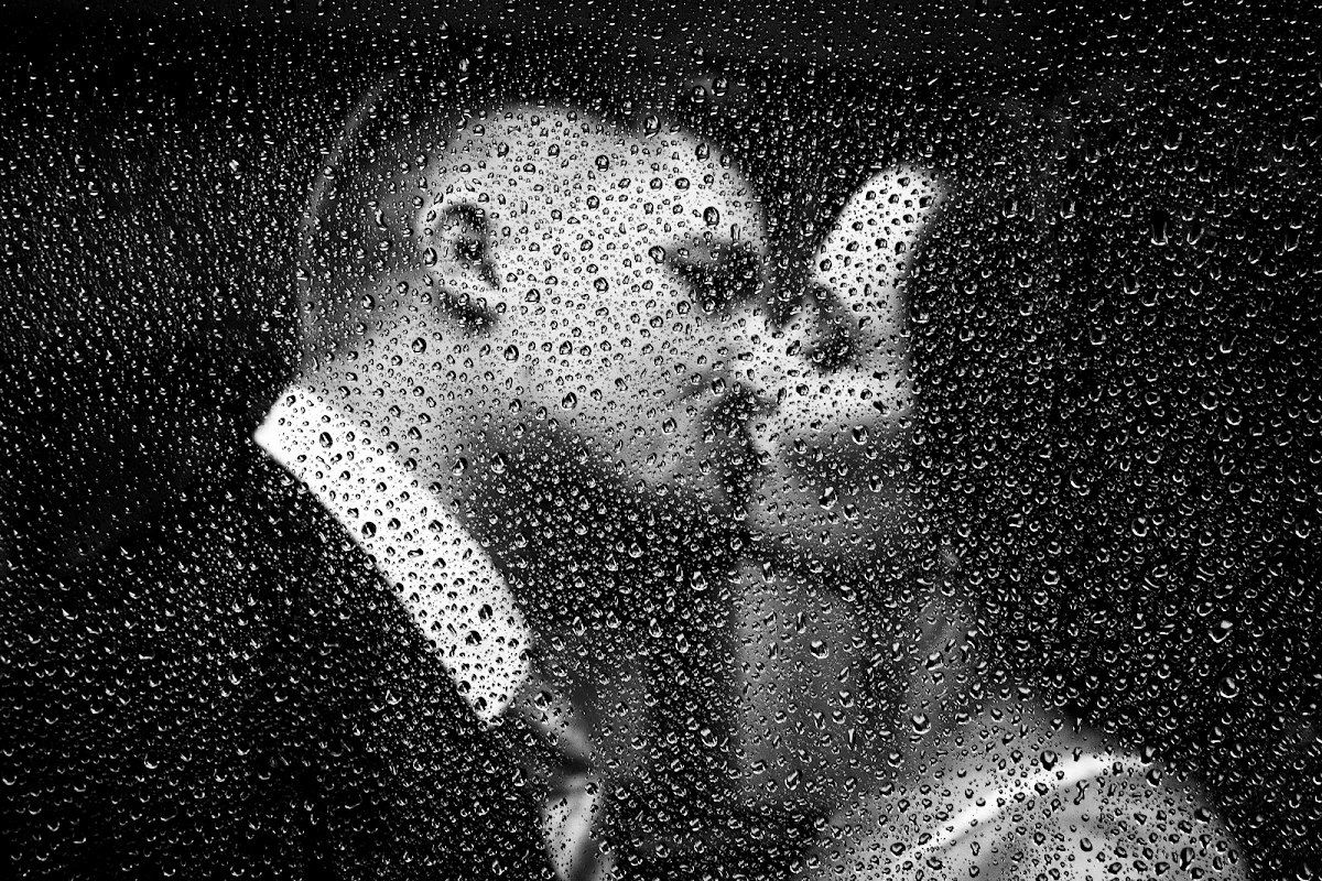 Retrato en blanco y negro de unos novios besándose tras una ventana cubierta de gotas de lluvia