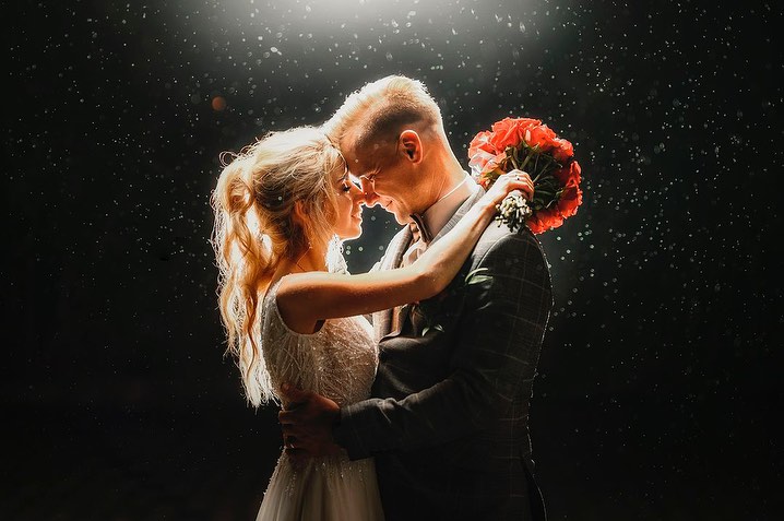 Novios abrazados bajo la lluvia iluminada de noche, con un ramo rojo detrás de la cabeza de él