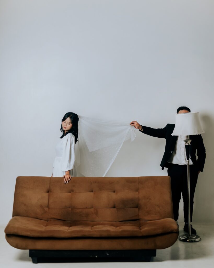 Couple behind a brown sofa, with the groom holding a veil and a lampshade partly obscuring his face