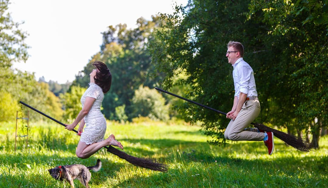 Pareja saltando con escobas en un jardín soleado, con un perro corriendo debajo.