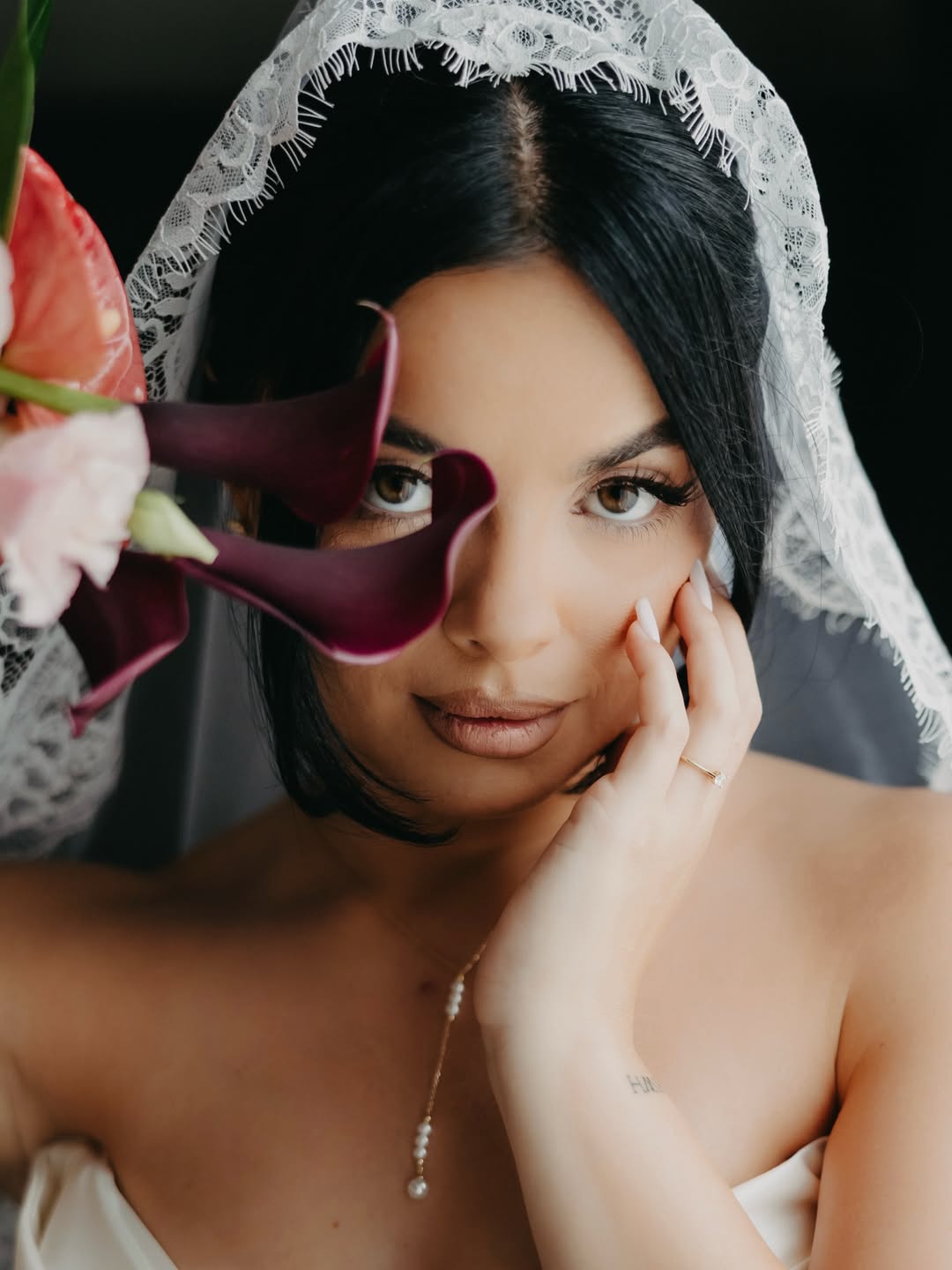 Bride in a lace veil with dark flowers beside her face and a hand against her cheek