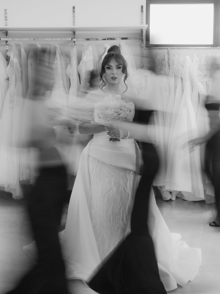 Black and white bridal portrait with a still face surrounded by blurred figures, photographed by Sabina Mladin