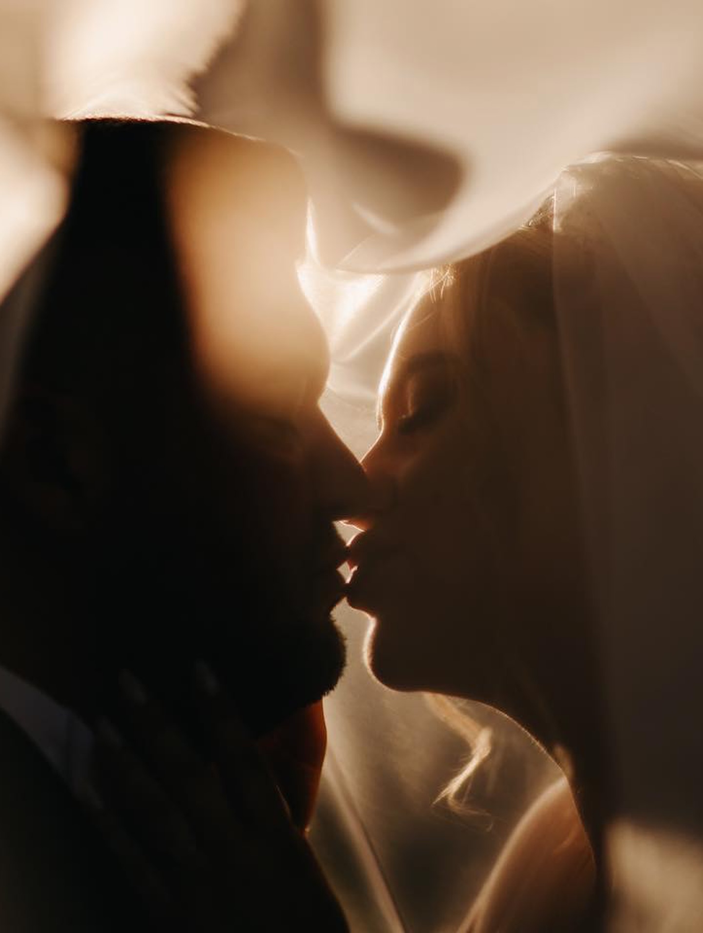 Bride and groom kissing beneath a softly backlit veil