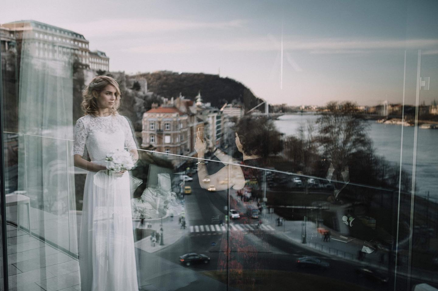 Novia con un ramo junto a una ventana, entre reflejos de la ciudad y el río.