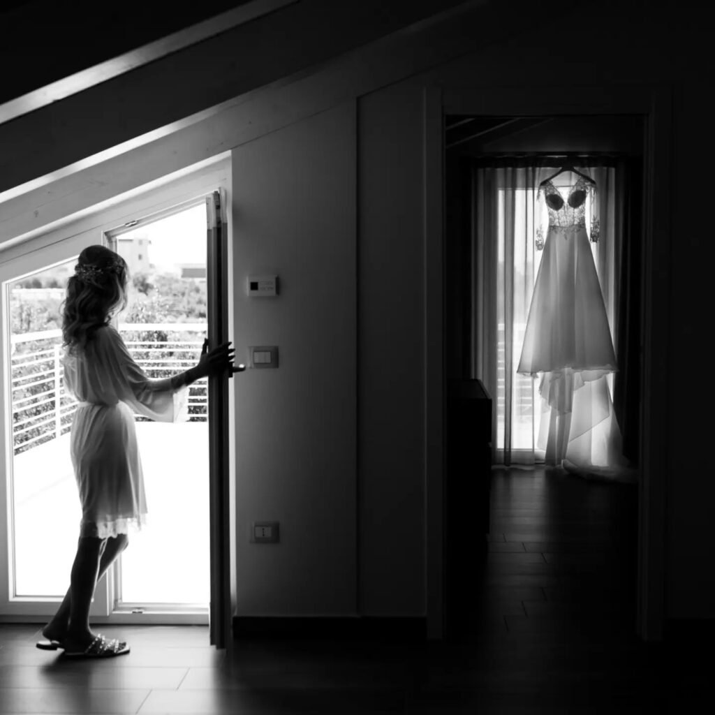 Black and white photograph of a bride by a window with her dress hanging nearby