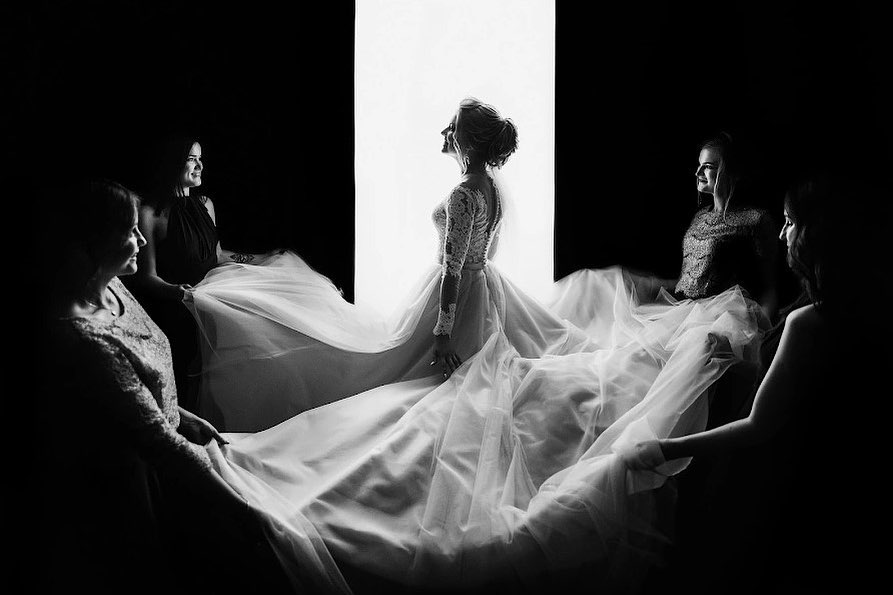 Bride and attendants arranging a flowing dress beside a bright window in black and white