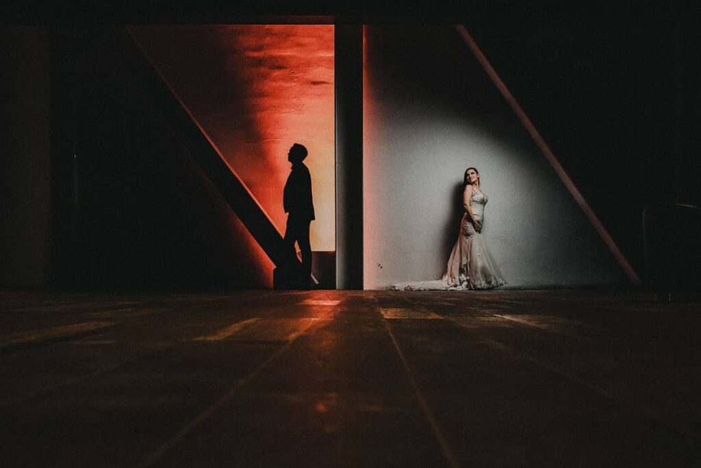 Novia iluminada de blanco y novio en una puerta roja, separados por la arquitectura oscura
