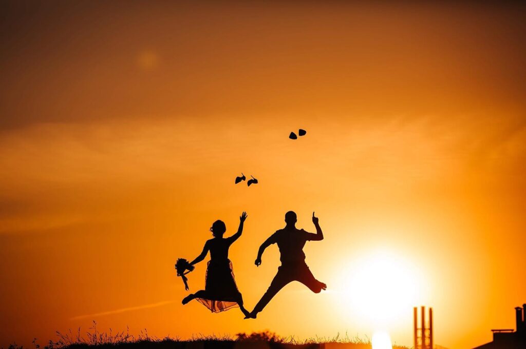 Couple holding hands and jumping in silhouette against an orange sunset