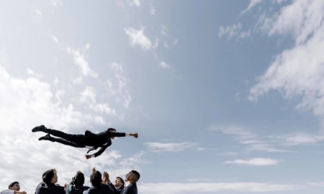 Groom airborne above guests' outstretched hands beneath a blue sky