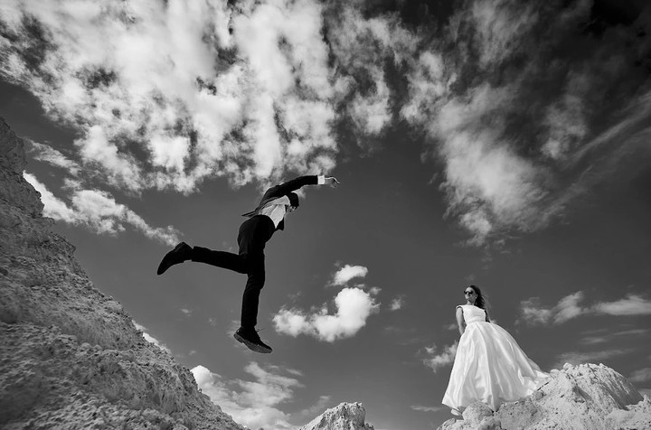 Groom leaping beside a bride beneath dramatic clouds in black and white