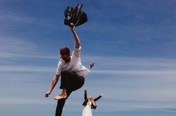 Barefoot groom jumping with his jacket raised and the bride behind him