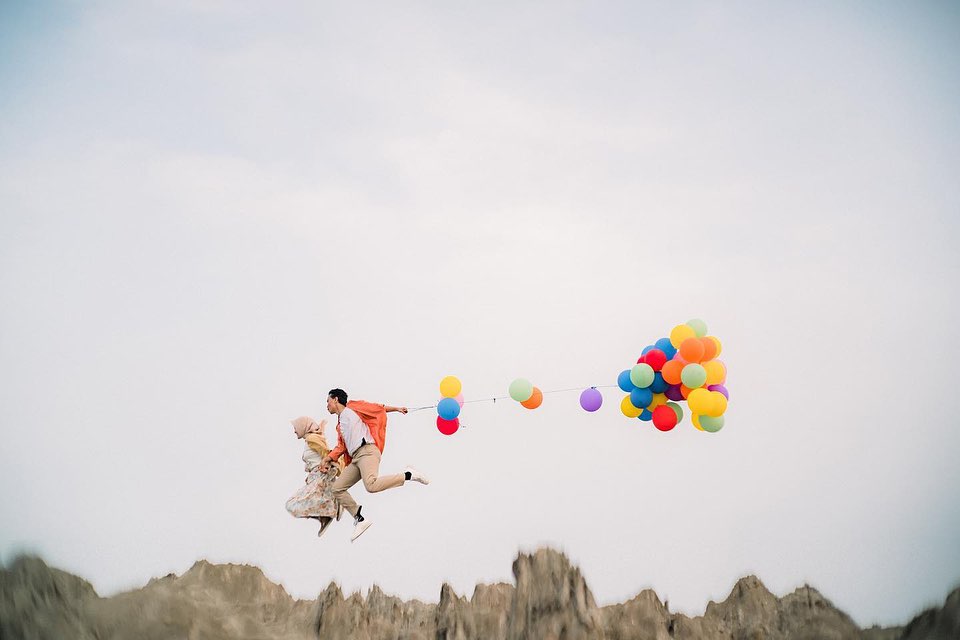 Couple airborne with colorful balloons trailing across a pale sky