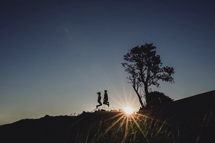Small couple silhouettes jumping above a ridge beside a sunburst