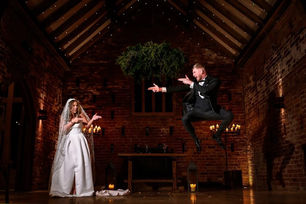 Groom jumping with crossed legs while the bride gestures inside a brick chapel