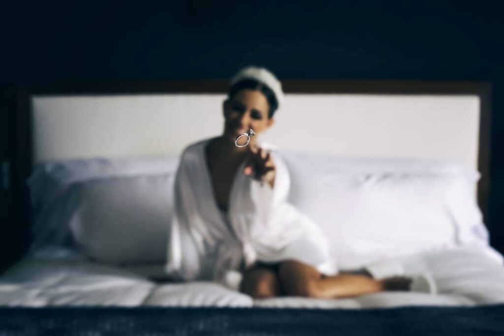 Bride holding a ring toward the camera while sitting on a bed