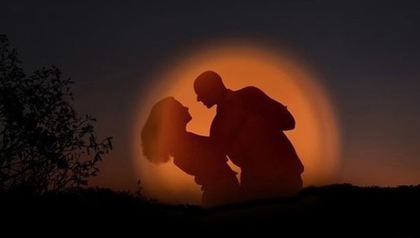 Couple leaning together in silhouette against an orange halo at dusk