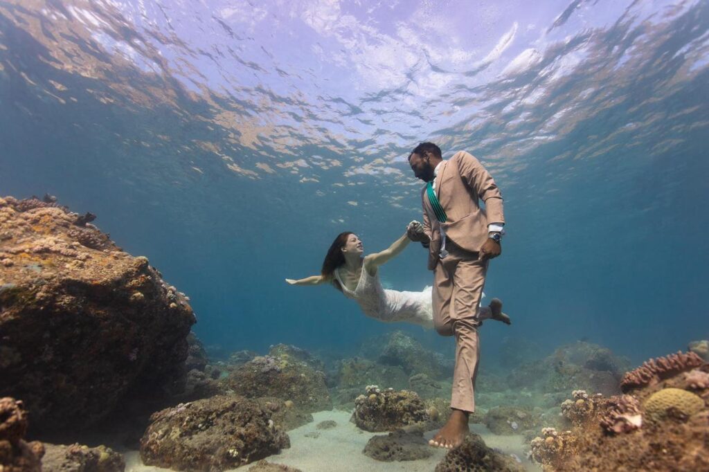 Wedding couple holding hands underwater between rocks beneath the surface