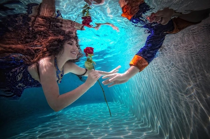 Underwater couple holding a red rose beneath bright blue reflections