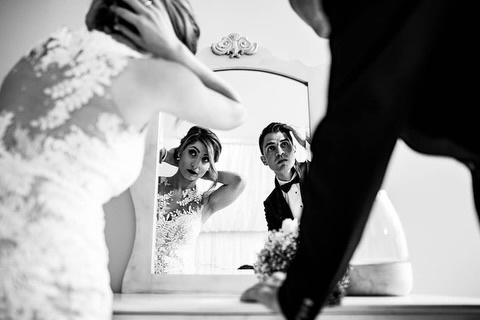 Black and white mirror view of a bride and man framed by fabric and nearby figures