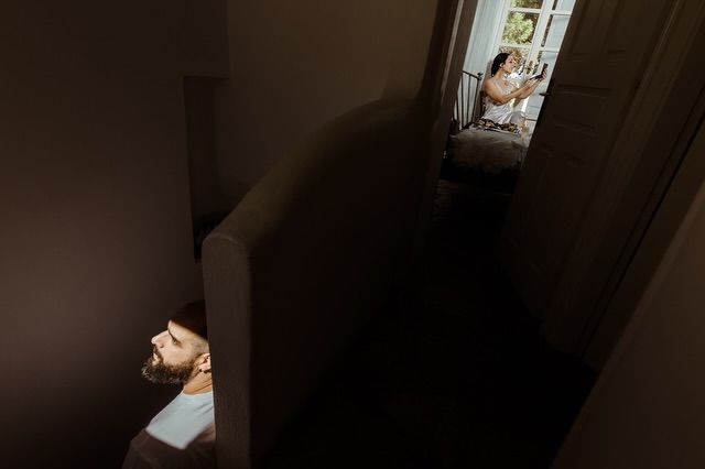 Layered view through a doorway showing a groom in the foreground and a bride at a vanity