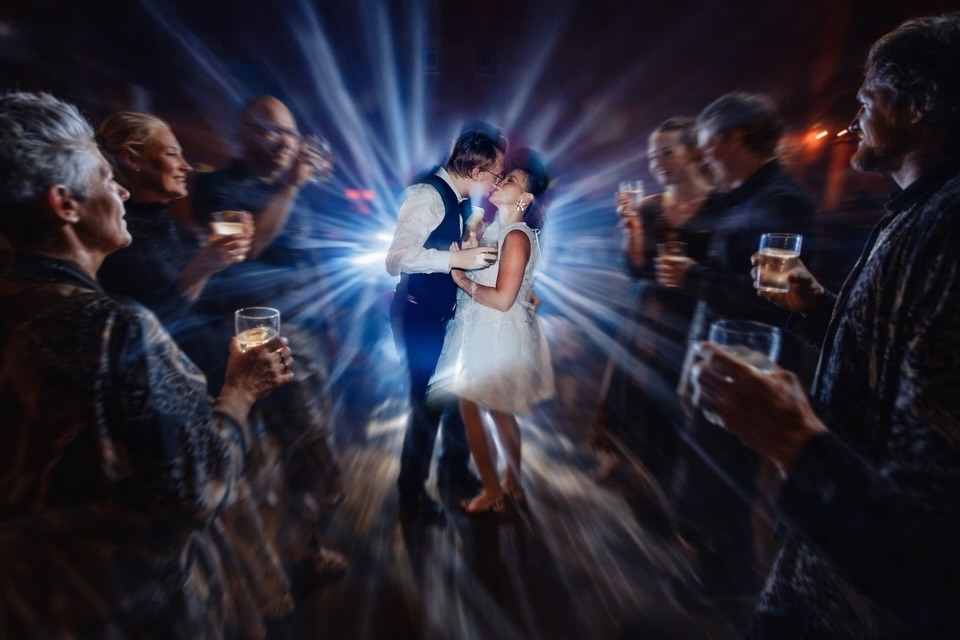 Couple kissing among guests holding drinks, surrounded by blue light streaks