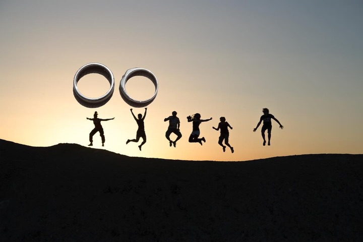 Grupo de siluetas saltando al atardecer bajo dos grandes formas de anillos