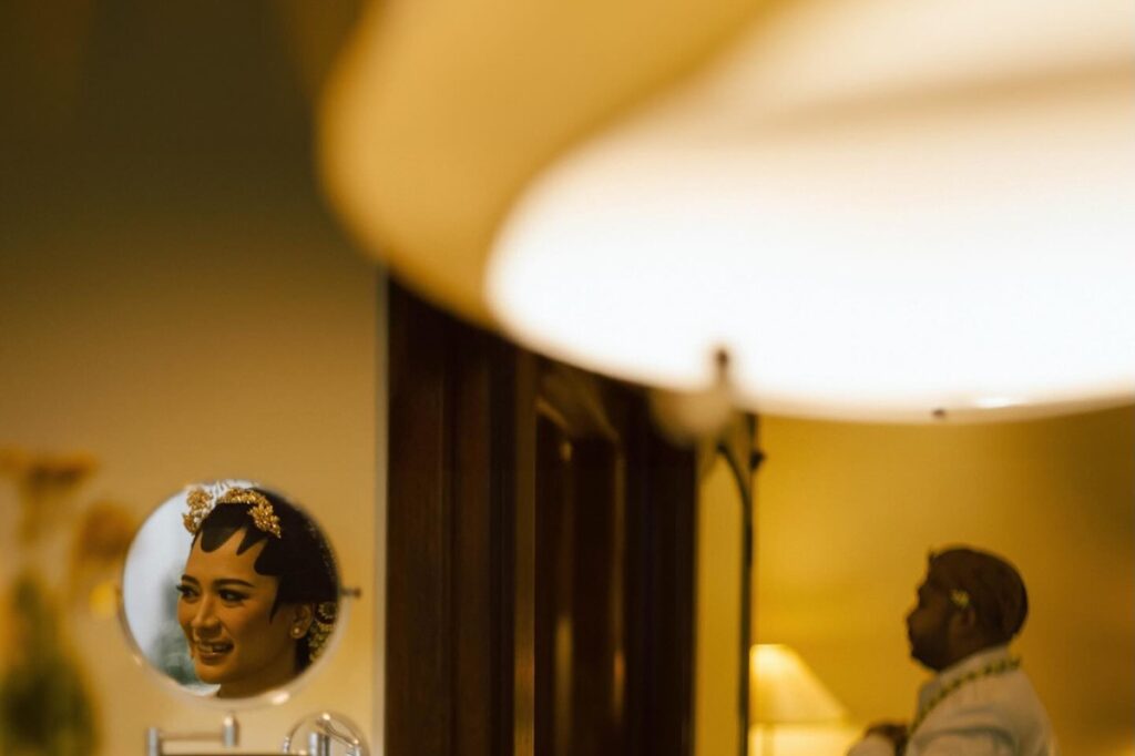 Bride reflected in a small mirror while a man sits in a warmly lit preparation room