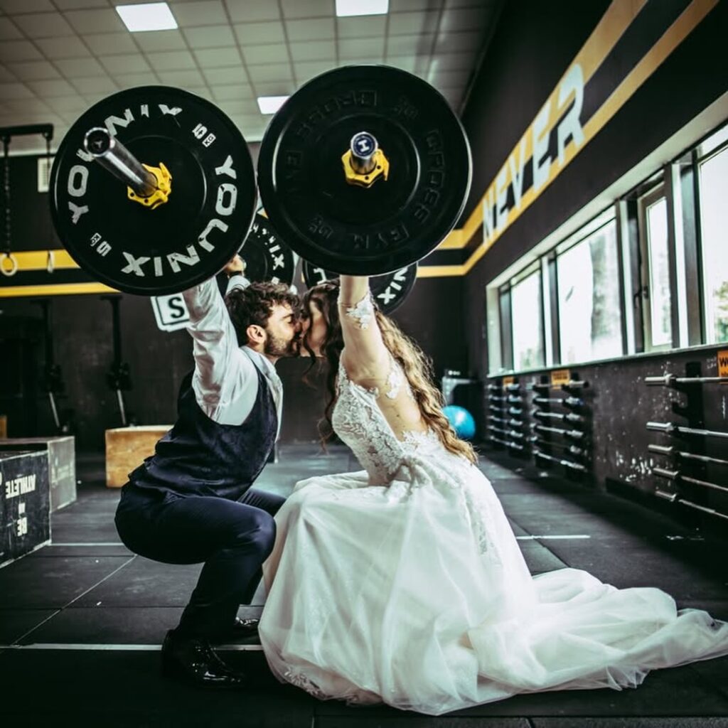 Pareja de boda besándose mientras sostiene barras con pesas en un gimnasio