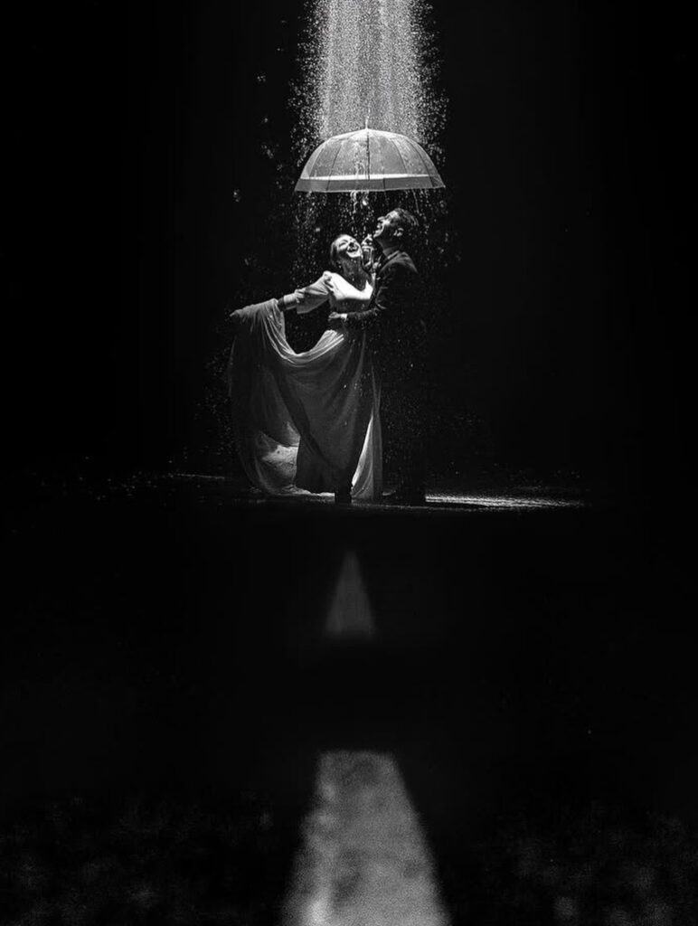 Retrato de boda en blanco y negro bajo un paraguas transparente, con lluvia iluminada cayendo alrededor