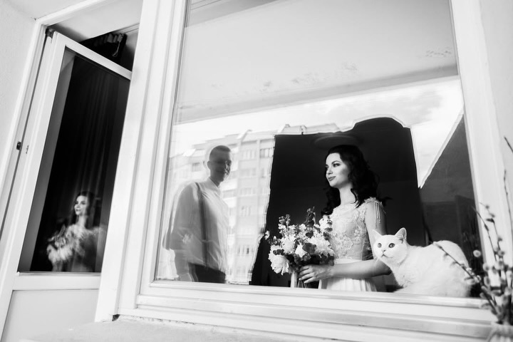 Black and white portrait of a bride at a window with a second figure visible in the glass