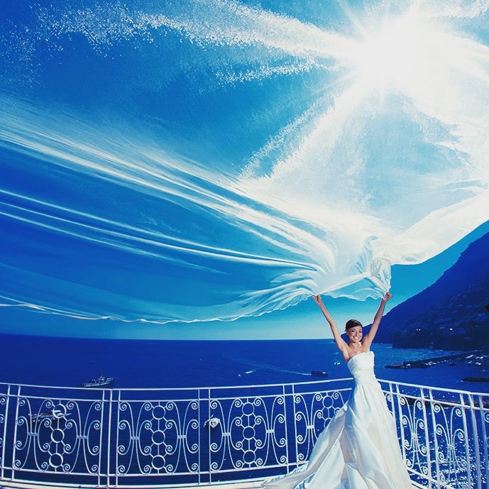 Bride holding a long white veil above her beside blue water