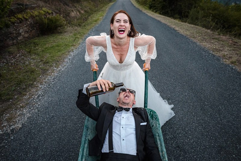 Novia riendo mientras empuja al novio en una carretilla por una carretera tranquila