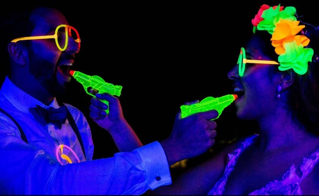 Retrato juguetón de una pareja con gafas, flores y pistolas de agua fluorescentes bajo luz azul