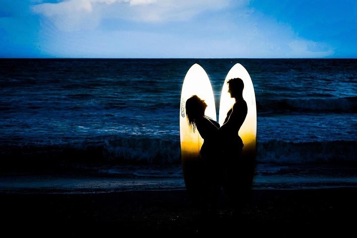 Couple in silhouette in front of two illuminated surfboards beside the sea at dusk