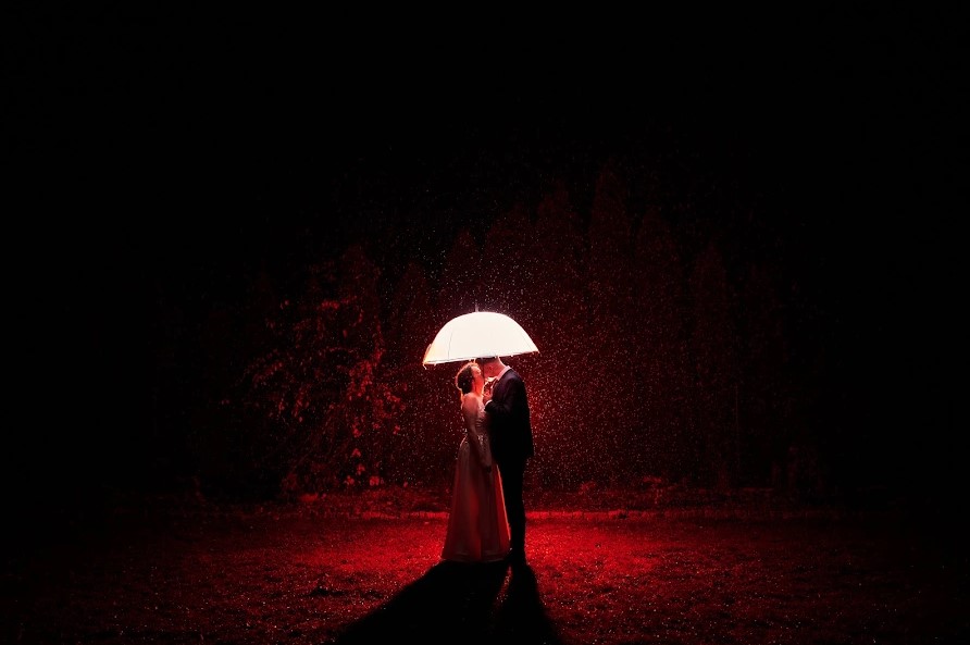 Novios bajo un paraguas con la lluvia iluminada en rojo contra la noche