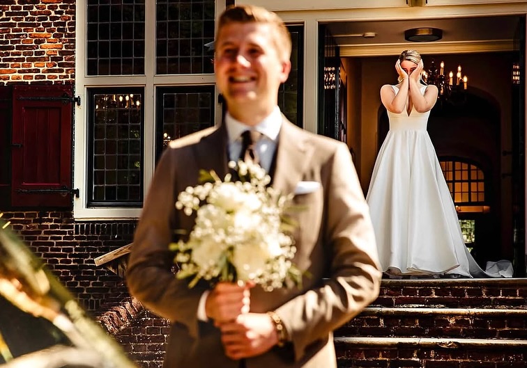 Novio sonriente con un ramo mientras la novia se cubre el rostro en una puerta