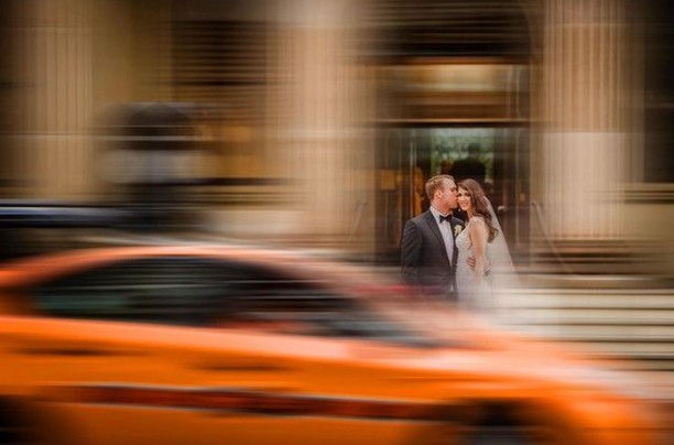 Una pareja de boda permanece visible detrás de un coche naranja que cruza desenfocado el primer plano