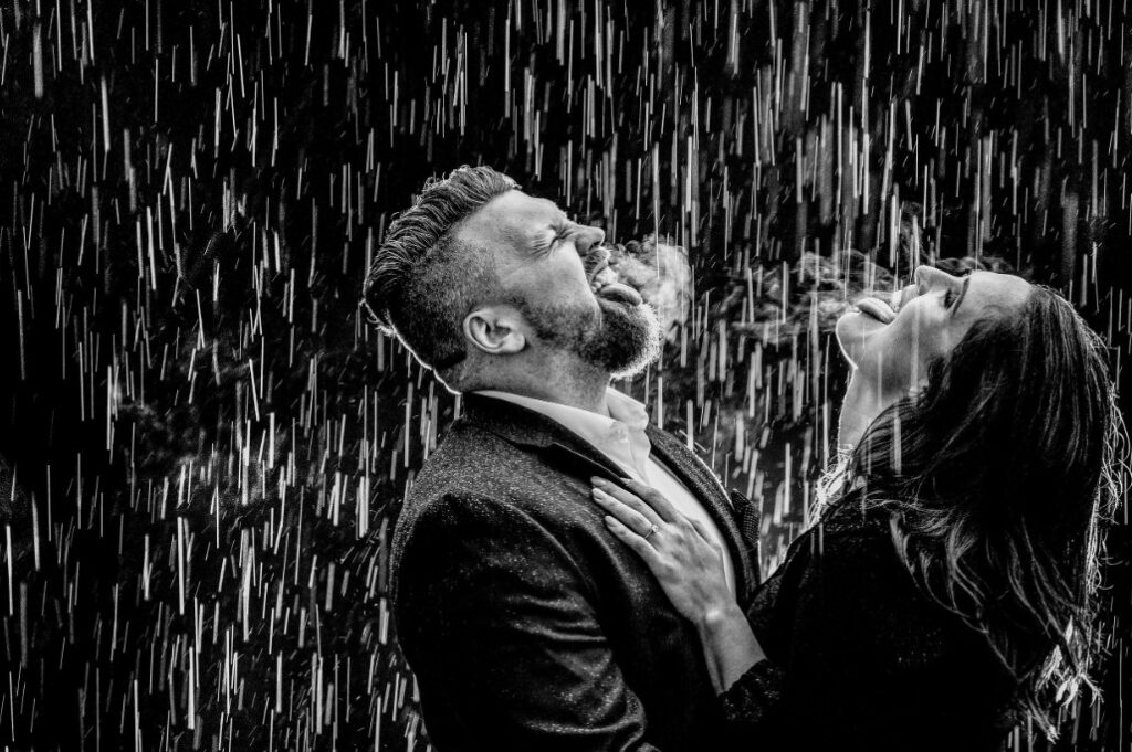 Fotografía en blanco y negro de una pareja riendo con el rostro hacia arriba bajo una lluvia intensa