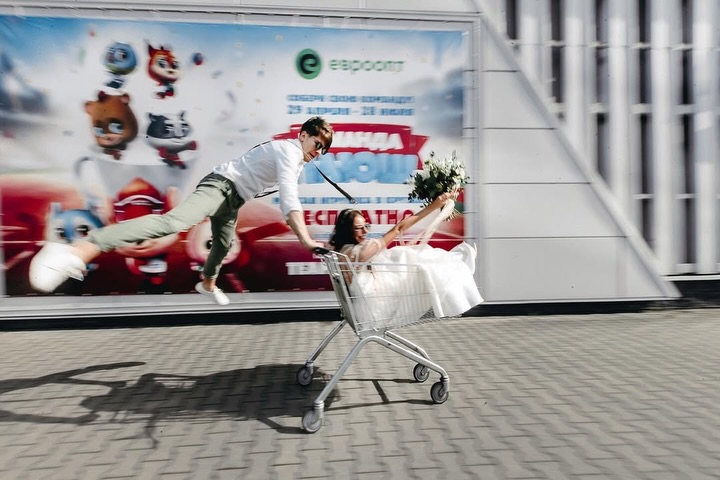 Novia en un carrito de compras junto al novio saltando en una calle