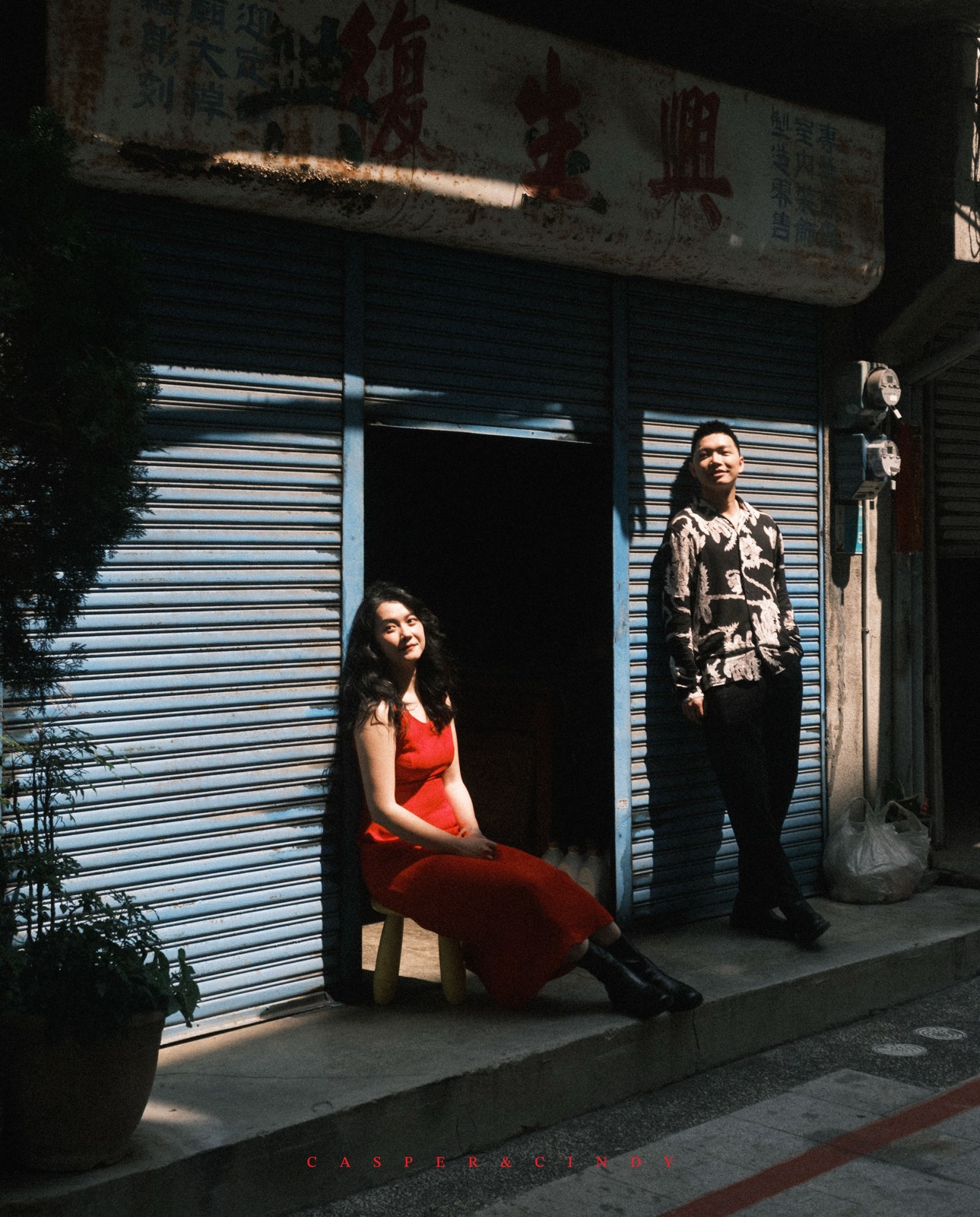 Pareja en la entrada soleada de un local con contraventanas azules y vestido rojo