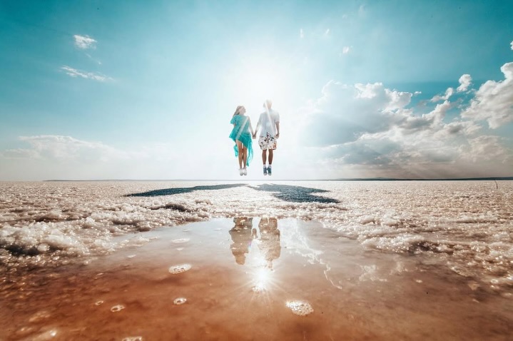Couple jumping in an open landscape with their reflections and the sun in a foreground puddle