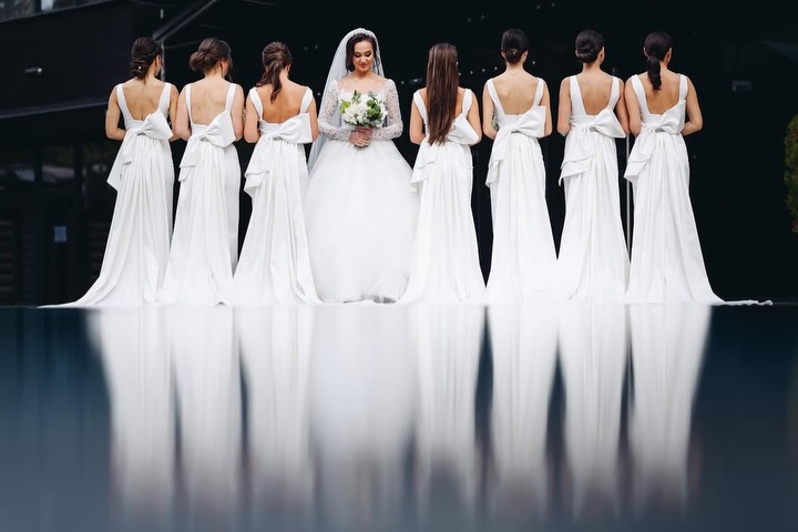 Novia mirando a cámara entre mujeres vestidas de blanco, con los vestidos reflejados debajo