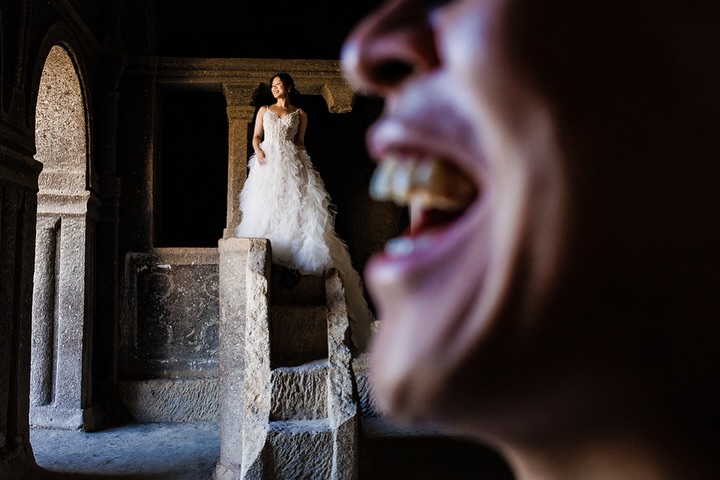 A smiling man close to the camera with a bride standing farther away inside a stone building