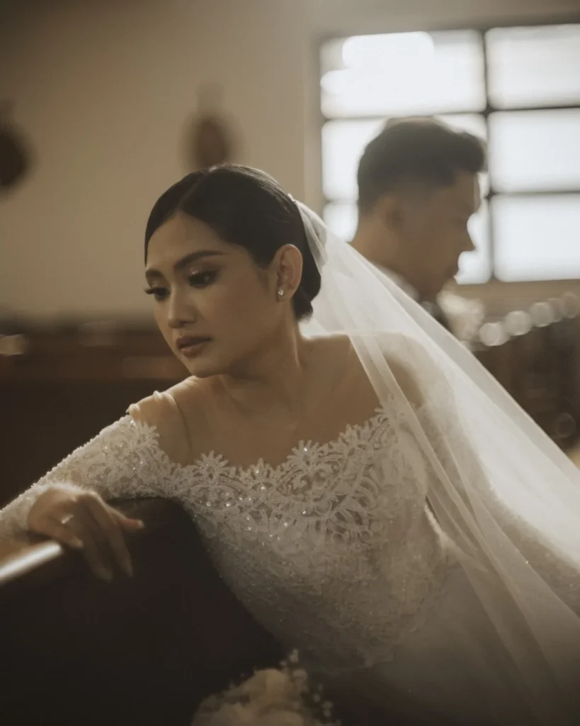 Bride in the foreground with her partner behind her near a window